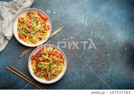 Two bowls with Chow Mein or Lo Mein, traditional Chinese stir fry noodles with meat and vegetables, served with chopsticks top view on rustic blue concrete background, space for text 107537383