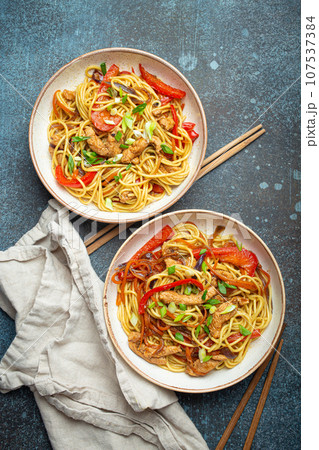Two bowls with Chow Mein or Lo Mein, traditional Chinese stir fry noodles with meat and vegetables, served with chopsticks top view on rustic blue concrete background 107537384