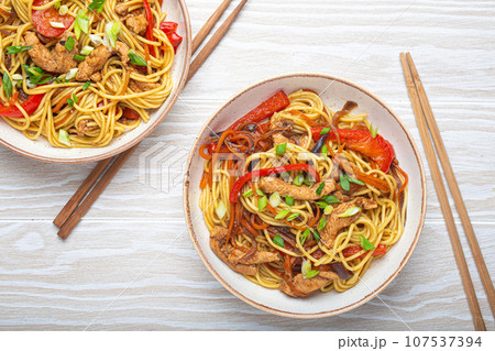 Two bowls with Chow Mein or Lo Mein, traditional Chinese stir fry noodles with meat and vegetables, served with chopsticks top view on rustic white wooden background table 107537394