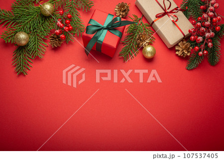 Christmas or New Year celebration red paper festive background with decoration fir tree, wrapped present boxes, cones, berries, sparkly red balls. Space for text. 107537405
