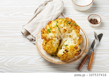 Whole roasted head of cauliflower with olive oil on plate on white wooden rustic table top view with cutlery. Vegetarian dish, space for text. Whole roasted head of cauliflower with olive oil on plate on white wooden rustic table top view with cutlery. Vegetarian dish, space for text. 107537418