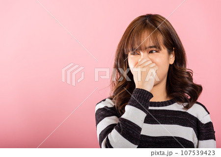 An unhappy Asian woman squeezes her nose in disgust due to a foul smell. Studio shot isolated on pink background, reflecting the concept of healthcare. An unhappy Asian woman squeezes her nose in disgust due to a foul smell. Studio shot isolated on pink background, reflecting the concept of healthcare. 107539423
