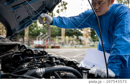man mechanical specialist professional checking oil level in car engine with tool on the road. Maintenance, safety, open supported hood auto. Side view of mechanic checking engine oil with open hood man mechanical specialist professional checking oil level in car engine with tool on the road. Maintenance, safety, open supported hood auto. Side view of mechanic checking engine oil with open hood 107543573
