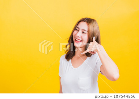 Portrait Asian Thai beautiful happy young woman wear white t-shirt standing smiling white teeth, making call me gesture by finger hand isolated on yellow background, with copy space Portrait Asian Thai beautiful happy young woman wear white t-shirt standing smiling white teeth, making call me gesture by finger hand isolated on yellow background, with copy space 107544856