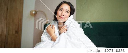 Beautiful romantic woman covers herself with warm duvet, enjoys sleeping and staying in bed, waking up happy in morning, sitting in bedroom 107548828