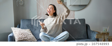 Portrait of carefree asian woman singing and listening music from smartphone app, using wireless headphones, smiling pleased, sitting on sofa at home Portrait of carefree asian woman singing and listening music from smartphone app, using wireless headphones, smiling pleased, sitting on sofa at home 107548993