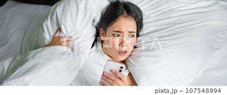 Close up portrait of asian girl lying in bed, looking at smartphone concerned, waking up late and staring at her alarm clock on mobile phone 107548994