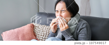 Close up of young korean woman staying at home with cold, sneezing in napking, has runny nose, concept of illness, health and influenza 107549017