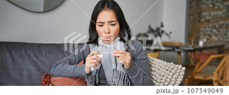 Shocked woman looks concerned at her thermometer, catching cold, has fever, sitting on sick leave at home Shocked woman looks concerned at her thermometer, catching cold, has fever, sitting on sick leave at home 107549049