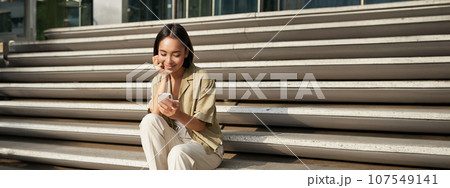 Vertical shot of asian woman, student sits on stairs in city, looking at mobile phone screen and smiling, using smartphone app Vertical shot of asian woman, student sits on stairs in city, looking at mobile phone screen and smiling, using smartphone app 107549141