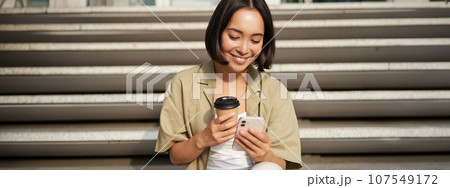 Happy urban girl drinks her takeaway coffee and scrolls feed on smartphone. Asian woman sits on stairs with tea and holding mobile phone Happy urban girl drinks her takeaway coffee and scrolls feed on smartphone. Asian woman sits on stairs with tea and holding mobile phone 107549172
