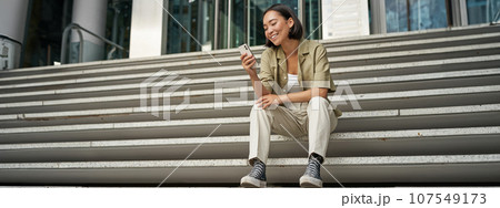 Portrait of happy smiling asian woman, sitting outdoors near building, using smartphone. Technology concept Portrait of happy smiling asian woman, sitting outdoors near building, using smartphone. Technology concept 107549173