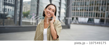 Smiling young korean girl talking on mobile phone and walking in city. Happy woman posing on street with smartphone Smiling young korean girl talking on mobile phone and walking in city. Happy woman posing on street with smartphone 107549175