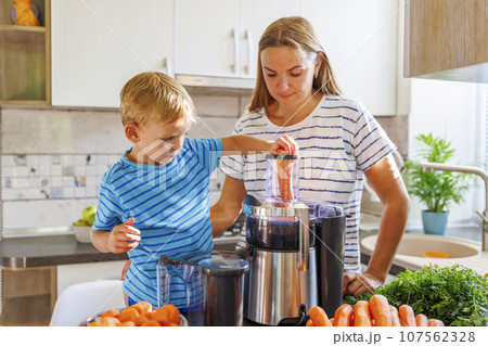 Little child boy with mother makes fresh carrot juice with juicer in kitchen Little child boy with mother makes fresh carrot juice with juicer in kitchen 107562328