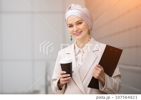 Muslim businesswoman walks with clipboard and carton cup Muslim businesswoman walks with clipboard and carton cup 107564821