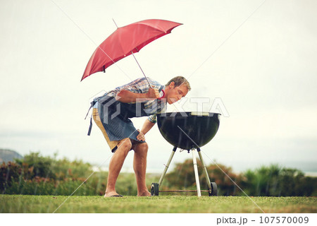 Umbrella, rain and a man outdoor to barbecue food for cooking or insurance in the winter season. Storm, weather and grill with a male person getting wet while trying to bbq on a grass lawn or garden 107570009
