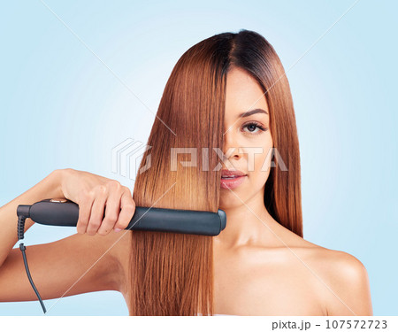 Face portrait, beauty and woman with hair straightener in studio isolated on a blue background. Haircare, product and female model with flat iron for salon treatment, hairstyle aesthetic or balayage. 107572723