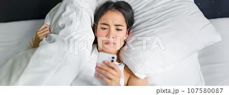 Close up portrait of asian girl lying in bed, looking at smartphone concerned, waking up late and staring at her alarm clock on mobile phone 107580087