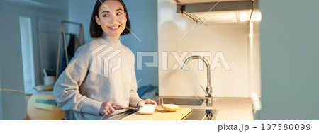 Cute asian woman making herself toast, cut loaf of bread, preparing sandwitch on kitchen Cute asian woman making herself toast, cut loaf of bread, preparing sandwitch on kitchen 107580099