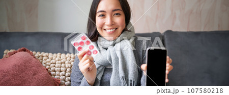 Smiling korean girl shows smartphone screen, medication in hands, feeling sick and staying at home, using online GP doctor app while on sick leave 107580218
