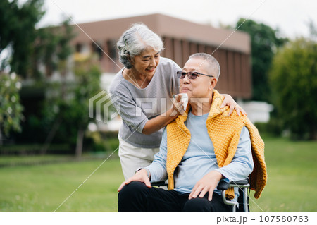 Asian Senior couple sitting in wheelchairs taking care of each other.in romantic time They laughing and smiling while sitting outdoor in park.. Asian Senior couple sitting in wheelchairs taking care of each other.in romantic time They laughing and smiling while sitting outdoor in park.. 107580763