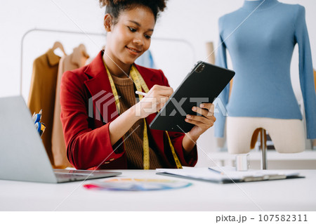Calm curly brunette dark skinned woman on desk in office of fashion designer and holds tablet and smartphone.. 107582311