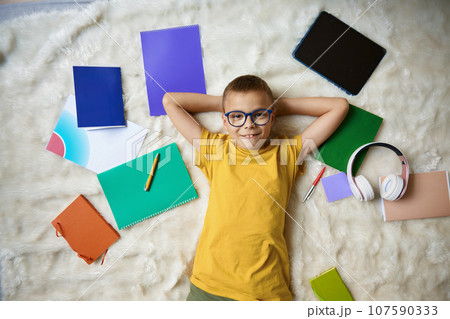 Happy smiling school boy rest in bed surrounded studying accessories Happy smiling school boy rest in bed surrounded studying accessories 107590333