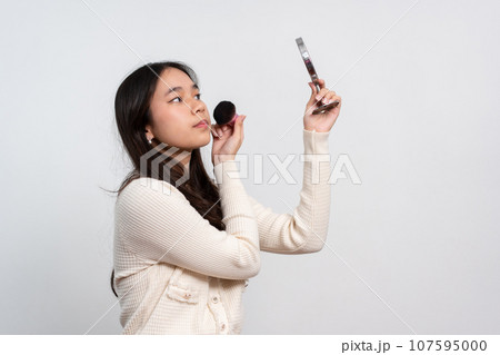 Beautiful young woman makes blush on her face using makeup brush on isolated white background. Teenage girl doing her own makeup. Close up portrait. Copy, empty space for text 107595000