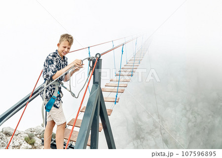 Boy fastens safety belt carabine to walk along rope bridge Boy fastens safety belt carabine to walk along rope bridge 107596895