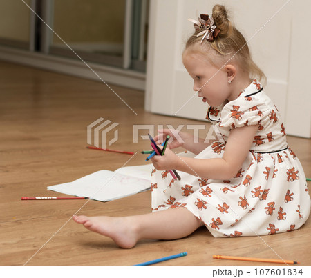 A little beautiful girl in a white dress sits on the floor in a home interior, draws and plays with colorful pencils. 107601834
