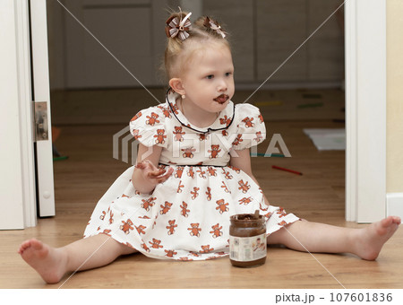 A little girl 3 years old in a beautiful white dress sits on the floor in a home interior and happily eats chocolate spread from a jar. His whole mouth is ridiculously smeared with chocolate. 107601836