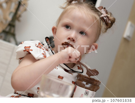 A little girl 3 years old in a beautiful white dress sits on the floor in a home interior and happily eats chocolate spread from a jar. His whole mouth is ridiculously smeared with chocolate. 107601838