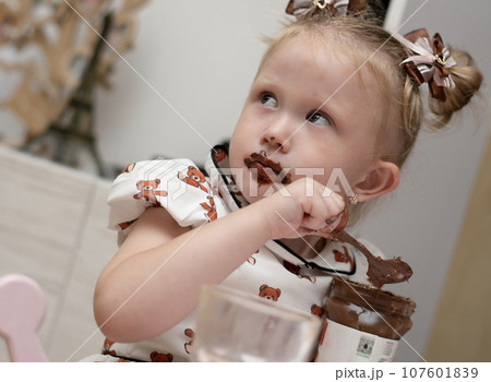 A little girl 3 years old in a beautiful white dress sits on the floor in a home interior and happily eats chocolate spread from a jar. His whole mouth is ridiculously smeared with chocolate. 107601839