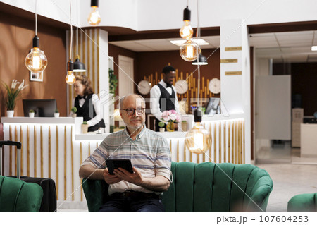 Caucasian senior person sits on hotel lobby sofa, holding digital tablet. Old man waiting in luxurious lounge area and uses smart device for browsing on internet. Luxurious and relaxing atmosphere. 107604253