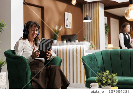 Elderly caucasian woman wearing headphones and sitting on sofa holding her tablet. Senior female guest having wireless headset waits in hotel lobby and uses digital gadget to browse the internet. Elderly caucasian woman wearing headphones and sitting on sofa holding her tablet. Senior female guest having wireless headset waits in hotel lobby and uses digital gadget to browse the internet. 107604282
