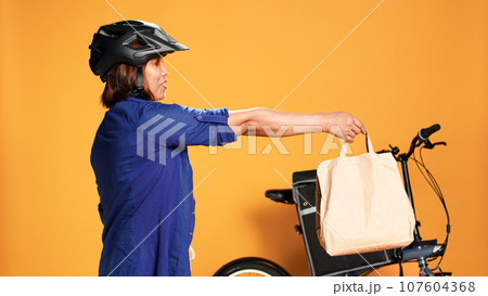 BIPOC food delivery worker reaching order address, waiting for customer to answer door. Courier woman greeting client, offering takeaway lunch bag, isolated over orange studio background 107604368