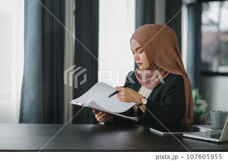 Young muslim millennial businesswoman in brown hijab headscarf working with financial reports while sitting in modern office. Young muslim millennial businesswoman in brown hijab headscarf working with financial reports while sitting in modern office. 107605955