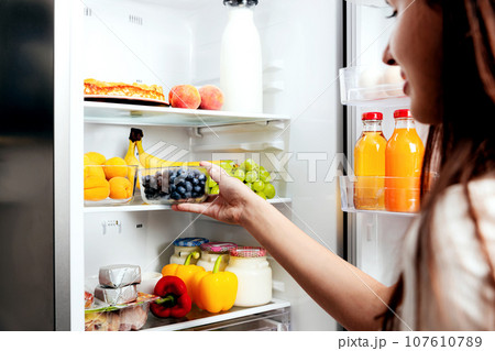 Woman hand taking, grabbing or picks up box of blueberry out of open refrigerator shelf or fridge drawer full of fruits, vegetables, banana, peaches, yogurt. Healthy food diet, lifestyle concept 107610789