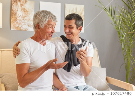 Laughing mature man sitting on couch with his adult son using smart phone together younger guy teaching his father how to use mobile phone and internet. Laughing mature man sitting on couch with his adult son using smart phone together younger guy teaching his father how to use mobile phone and internet. 107617490