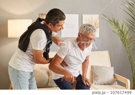 Caring son helping senior disabled man to stand up with walking stick, young man supporting orthopedic patients to get up with walking cane at home. International Day for the Elderly. 107617499