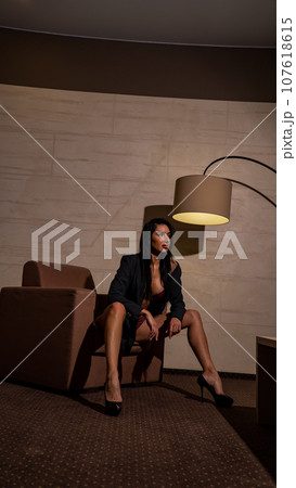Indoor shot of sexy young woman. Portrait of beautiful informal girl dressed in black underwear, leather jacket and sandals. Seductive lady sitting on a chair. Indoor shot of sexy young woman. Portrait of beautiful informal girl dressed in black underwear, leather jacket and sandals. Seductive lady sitting on a chair. 107618615