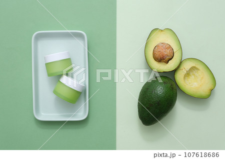Minimal scene with avocado ingredient and cosmetic jar unbranded for advertising. Top view of a halves of avocado on light green background and cream jars on plate on dark green background 107618686