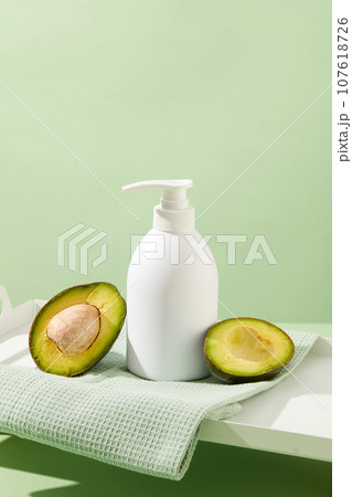 Minimalist concept for advertising cosmetic based on avocado extract. A white pump bottle placed on cotton towel with halves of avocado on pastel background. Empty label for design. Front view Minimalist concept for advertising cosmetic based on avocado extract. A white pump bottle placed on cotton towel with halves of avocado on pastel background. Empty label for design. Front view 107618726