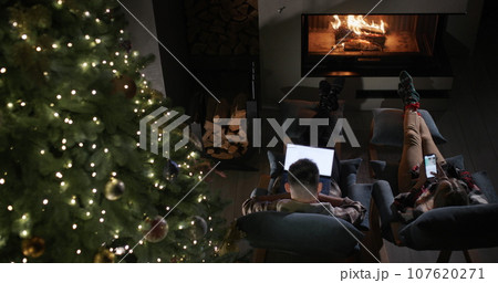 A young couple uses a smartphone and a laptop. Sitting by a tall Christmas tree and a fireplace A young couple uses a smartphone and a laptop. Sitting by a tall Christmas tree and a fireplace 107620271