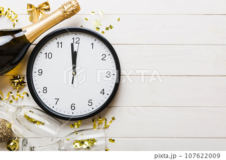 New year celebration concept with a bottle of champagne and two glasses toasting. Christmas gift box, alarm clock and fir tree branch on colored table. Top view Copy space 107622009
