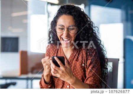 Young beautiful hispanic woman with curly hair inside office celebrating victory success, businesswoman received win notification on phone, using smartphone app at workplace. 107626877