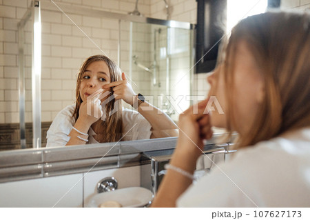 Cute teenage girl squeezes out a pimple on her cheek in the bathroom 107627173