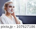 Young pretty girl with a white moisturizing mask applied to her face. A blue eyed blonde in a white robe does spa treatments in a beauty salon. 107627186