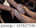 Close up of the masseuse palms with a grey scrub on them. Massage therapist applying cream to the female client legs. 107627190