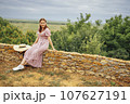 Beautiful young woman is sitting on stone fence and posing for camera. Charming girl in pink dress and wide brimmed straw hat smiles and rests against background of bushes and wide green meadows. 107627191